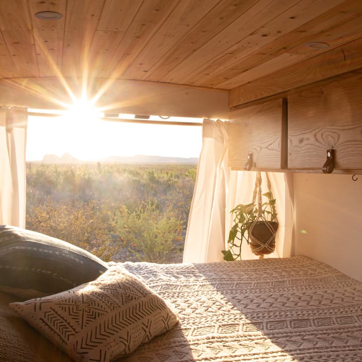 Cozy van interior with a bed, patterned pillows, wooden ceiling, and sunlight streaming through curtains.