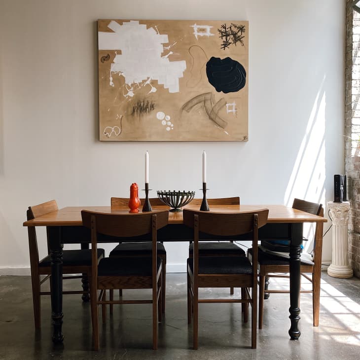 Dining room with wooden table, six chairs, abstract wall art, candlesticks, and a red pepper grinder.