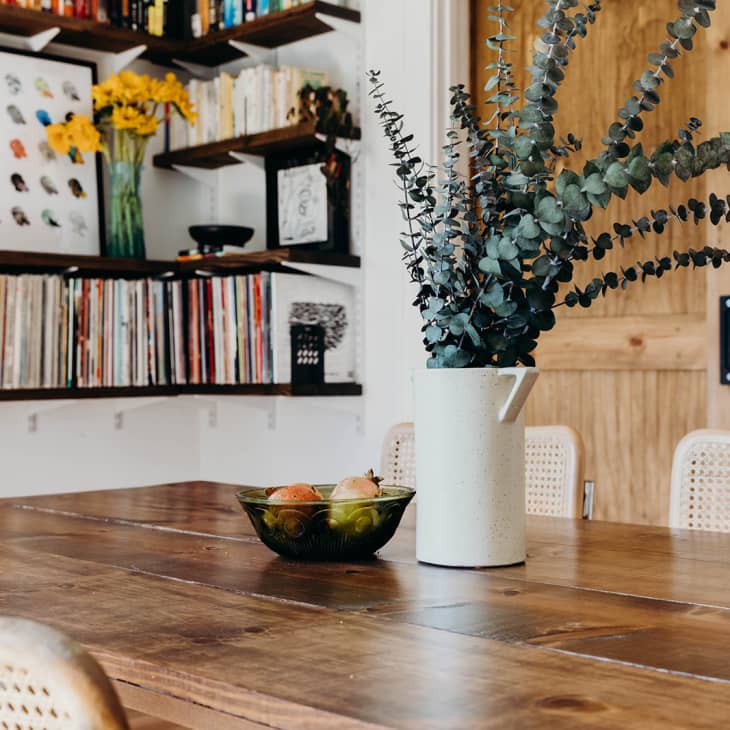 Dining table with eucalyptus in a white vase, fruit bowl, and wicker chairs, shelves with books and art in the background.