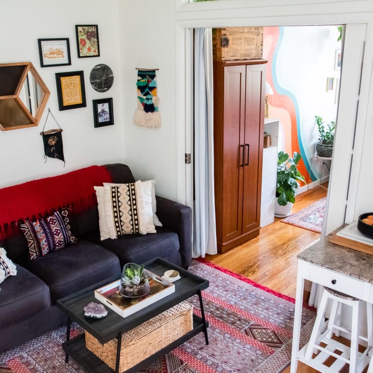 Cozy living room with a dark sofa, patterned pillows, wall art, and a small kitchen area with plants and wine bottles.