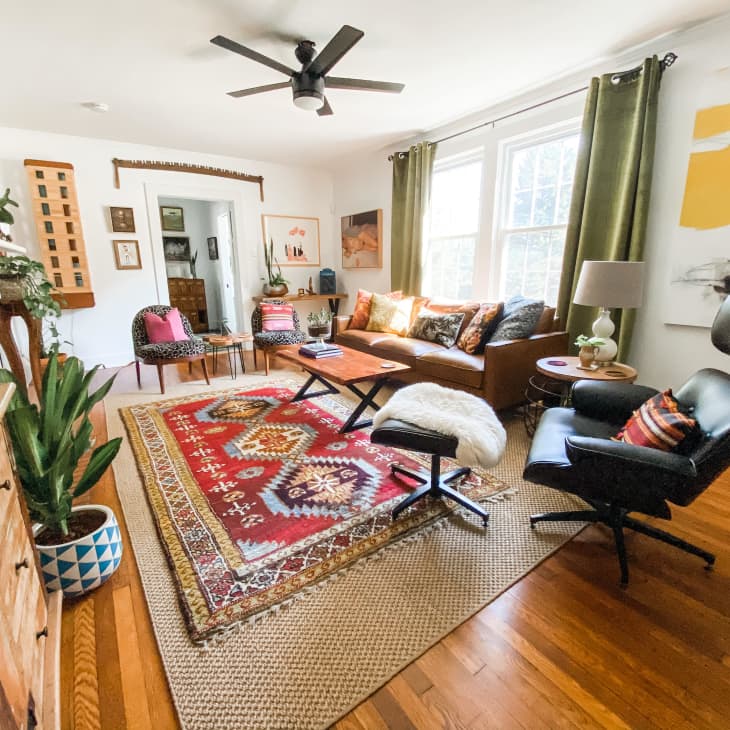 Living room with a brown sofa, colorful cushions, red patterned rug, black lounge chair, and green curtains.