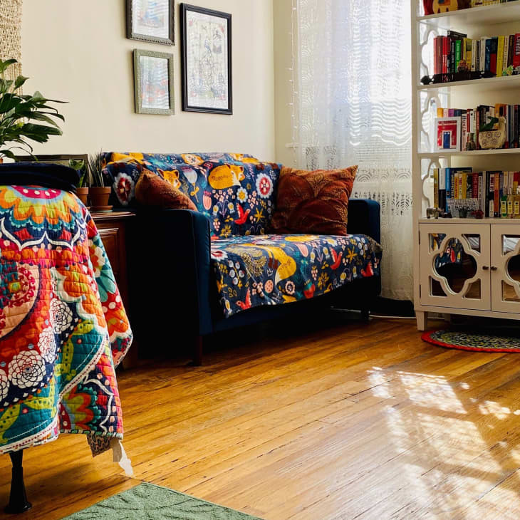Cozy living room with colorful quilted sofa, vibrant bedspread, potted plant, and white bookshelf filled with books.