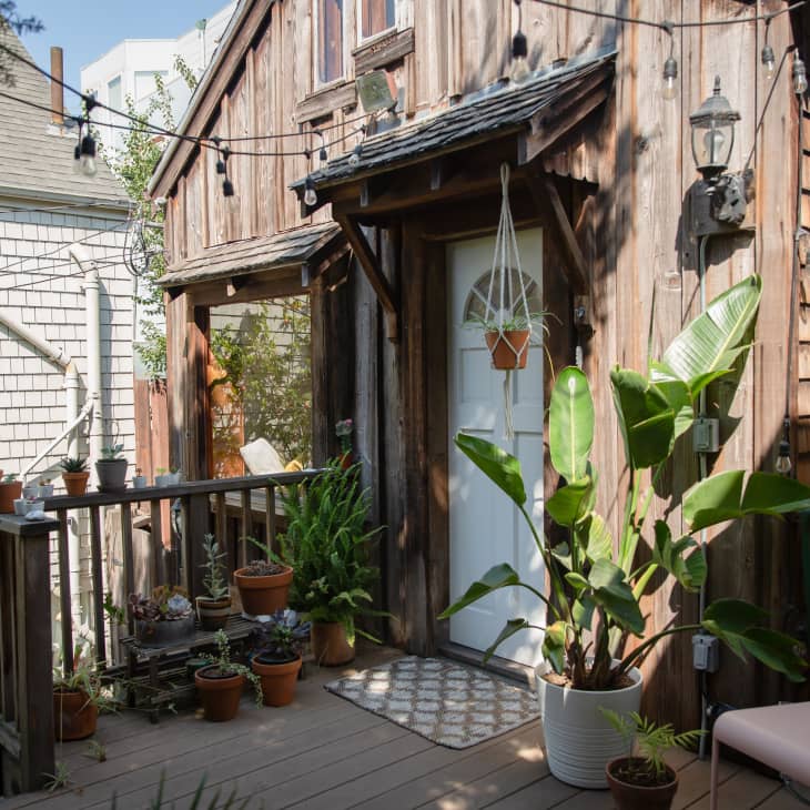 Rustic wooden cabin porch with potted plants, string lights, and a white door.