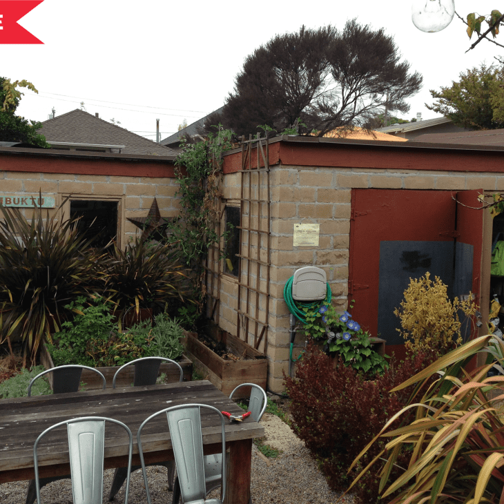 Outdoor patio with metal chairs, wooden table, lush plants, and a brick shed labeled "Timbuktu" with a star decoration.