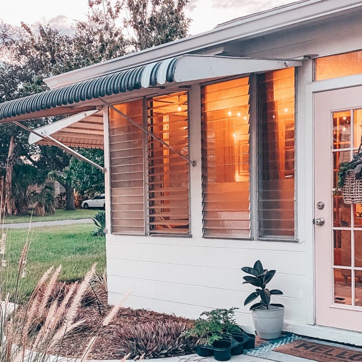 White house with striped awning, glass door, potted plants, and warm interior lighting.