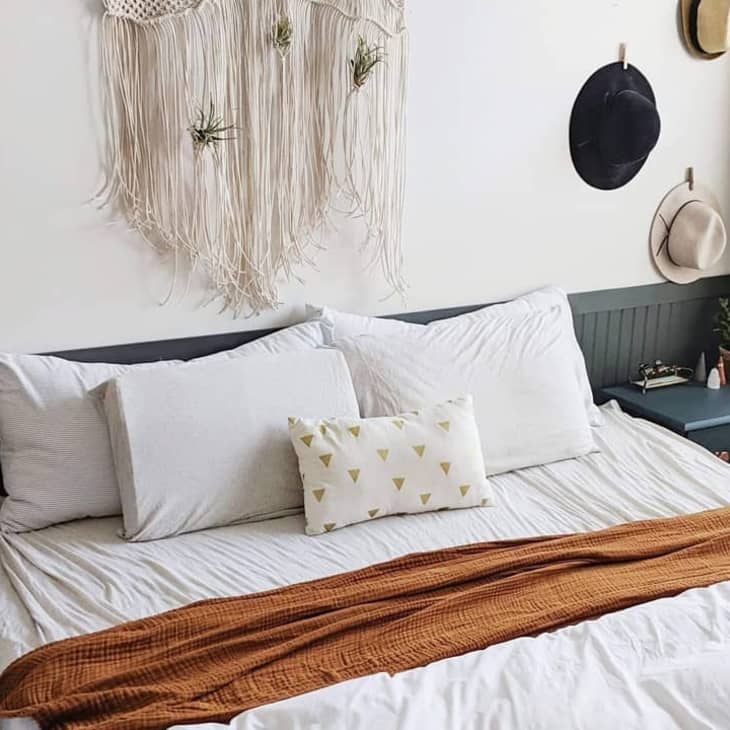 Bedroom with white bedding, a brown throw, macramé wall hanging, and hats on the wall.
