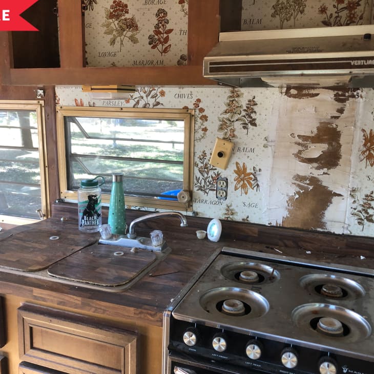 Vintage kitchen with wood cabinets, floral wallpaper, and a four-burner stove. Two windows above the sink.
