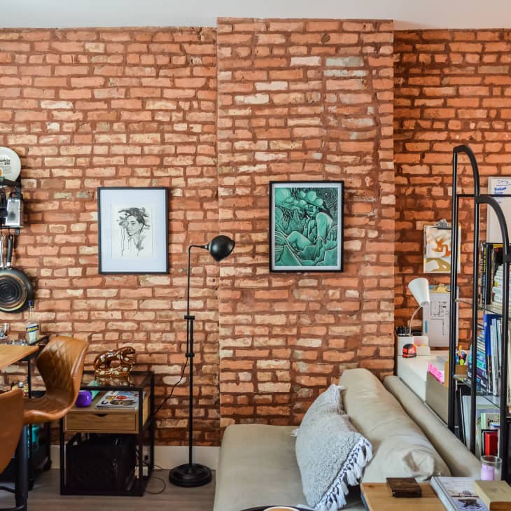 Exposed brick living room with wall art, open shelving, kitchenware, and a cozy seating area with a floor lamp.