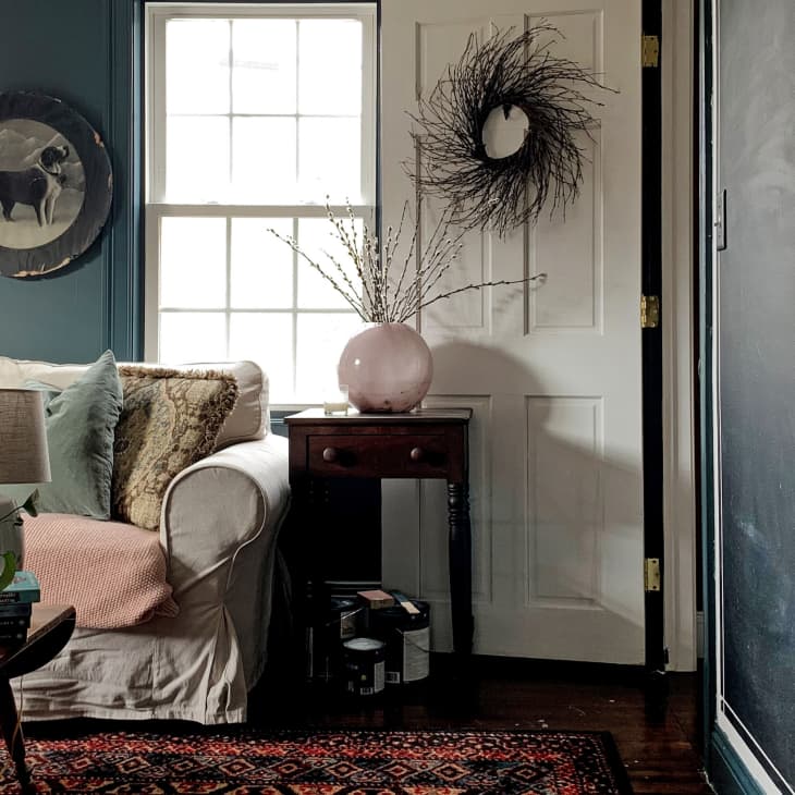 Cozy living room with a white sofa, green pillows, a plant, and a side table with a pink vase and wreath on the door.