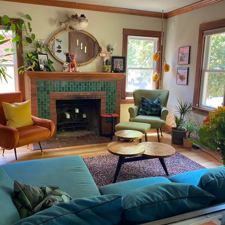 Living room with green-tiled fireplace, orange and green armchairs, blue sofa, plants, and colorful wall art.