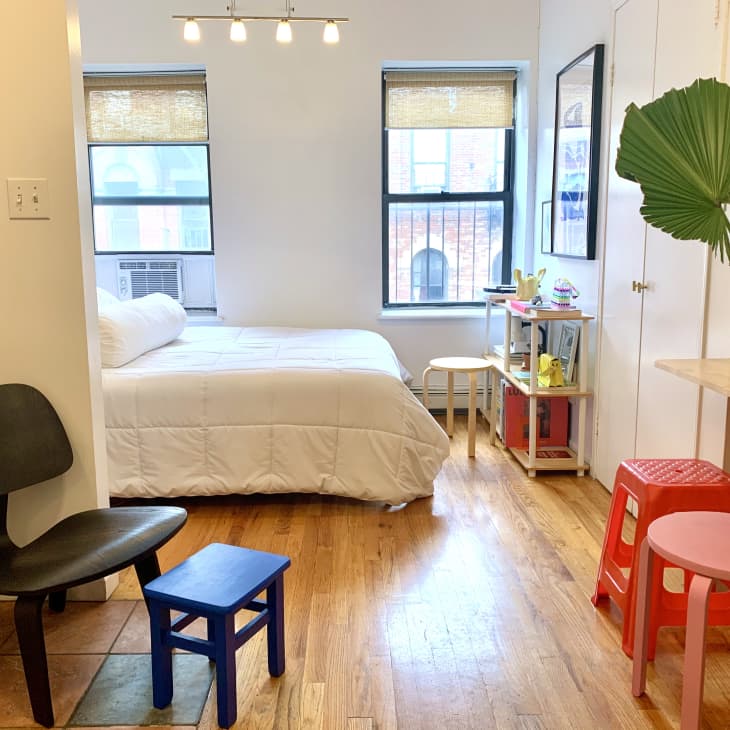Studio apartment with a bed, colorful stools, black chair, and large green leaves in a vase on a wooden floor.