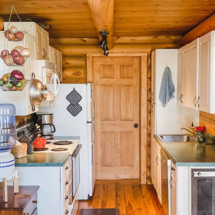 Rustic kitchen with log walls, hanging fruit baskets, water cooler, and wooden cabinets.