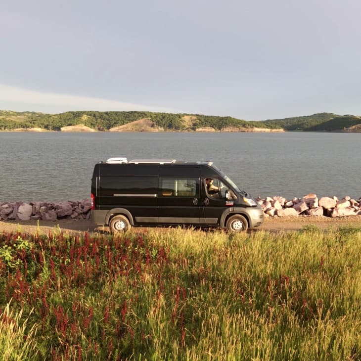 Black camper van parked by a lake with rocky shore, surrounded by grassy fields and distant hills.
