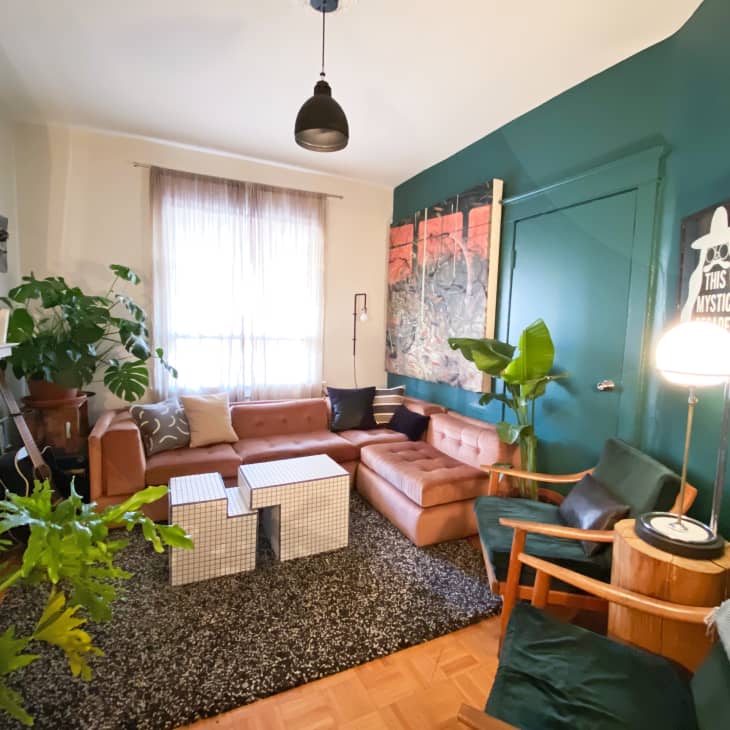 Cozy living room with brown sectional sofa, green accent wall, abstract art, plants, and modern lighting.