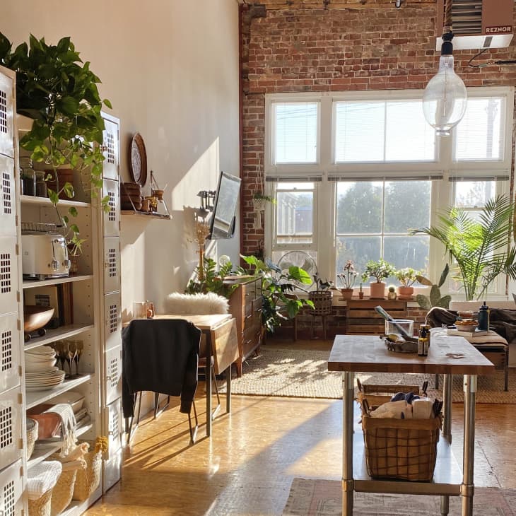 Loft kitchen with brick wall, large windows, plants, open shelving, and a central island table.