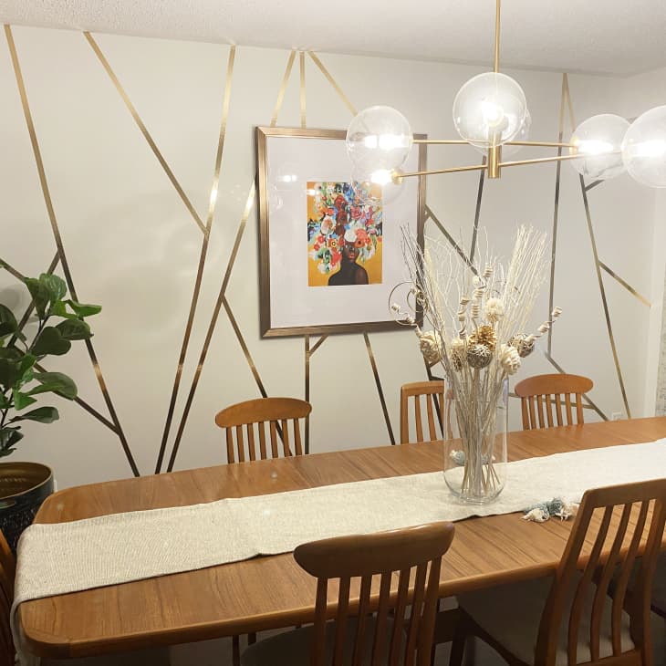 Dining room with wooden table, six chairs, geometric wall art, modern chandelier, and vase with dried branches.
