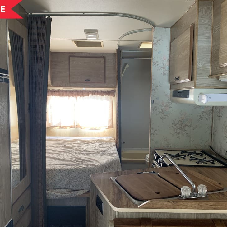Small RV interior with a kitchenette, stove, wooden cabinets, and a bed with floral curtains.