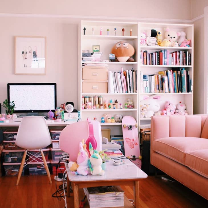 Home office with a pink sofa, white desk, computer, bookshelves filled with books, toys, and decor items.