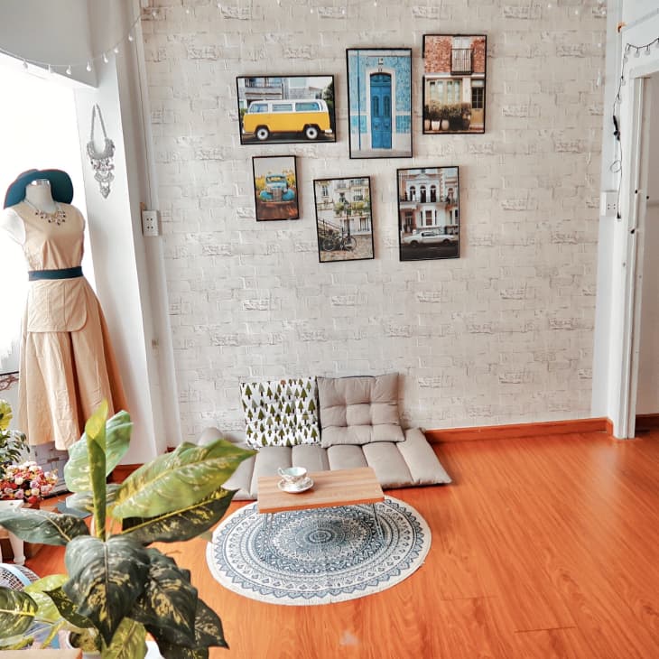 Cozy room with mannequin in beige dress, wall art, floor cushions, small table, and plants by a window.