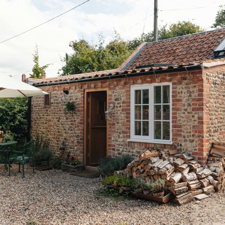 Stone cottage with a red-tiled roof, woodpile, and outdoor seating under a white umbrella in a garden setting.