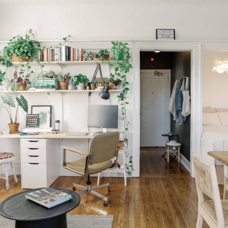Home office with white desk, plants, bookshelves, and a beige chair, adjacent to a cozy bedroom and dining area.