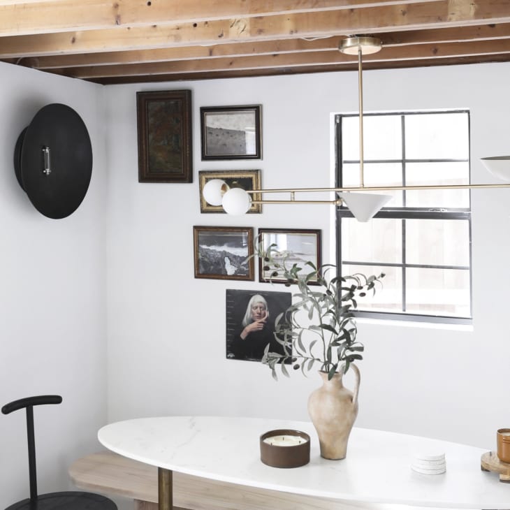Dining area with oval table, modern chandelier, framed art, and potted plant under exposed wooden beams.