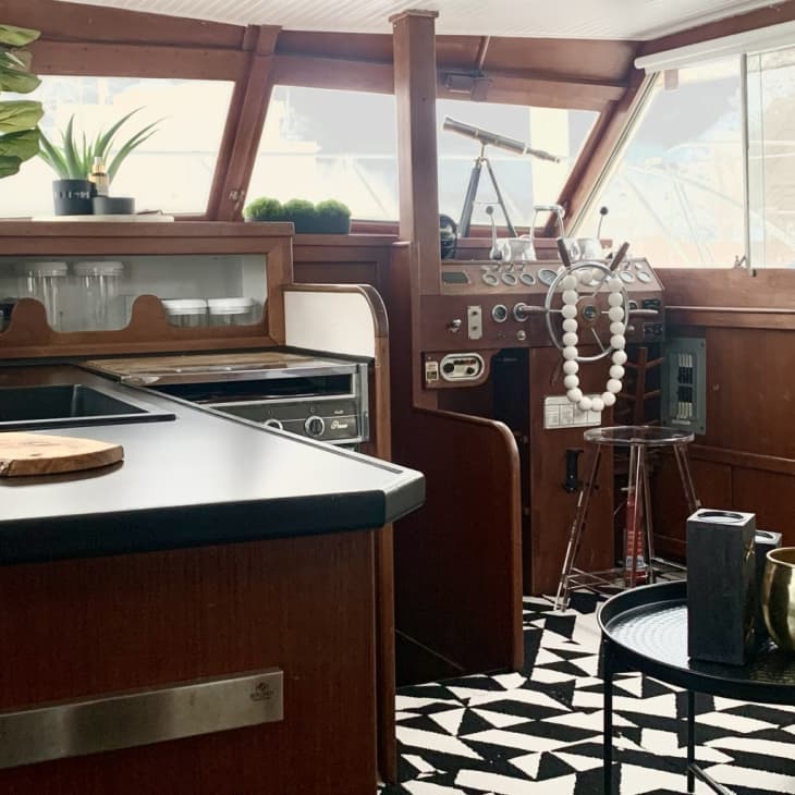 Boat interior with wooden helm, black countertop, cutting board, potted plants, and geometric patterned rug.