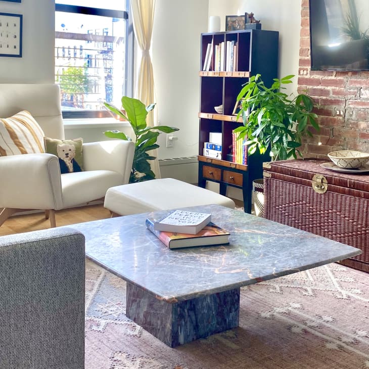 Cozy living room with a white armchair, marble coffee table, wicker chest, and plants against a brick wall.