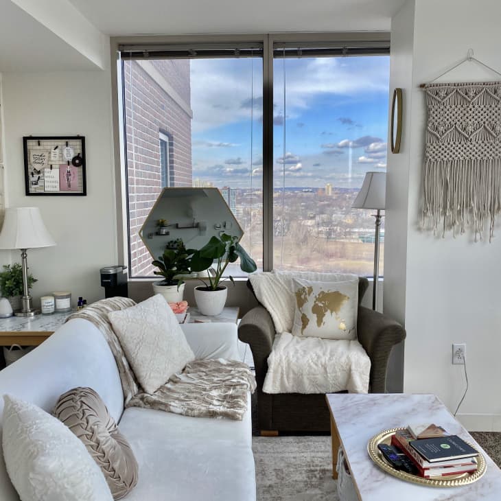 Cozy living room with a white sofa, brown armchair, macramé wall hanging, and large windows overlooking a cityscape.