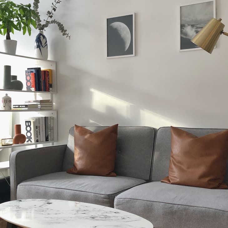 Gray sofa with brown leather pillows next to white bookshelf with plants on top