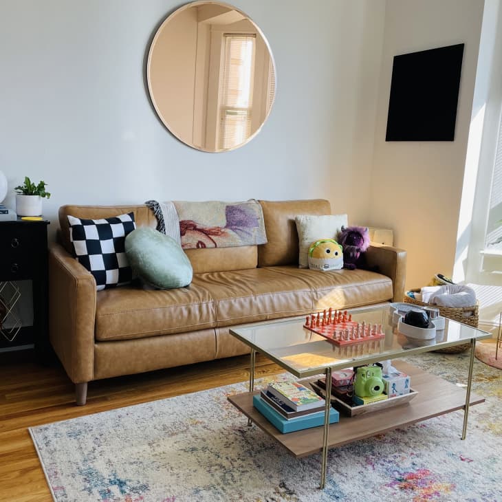 Living room with tan leather sofa, checkered pillow, glass coffee table with chess set, and round mirror on the wall.