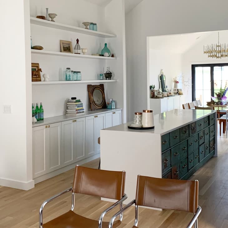 Open kitchen with white cabinets, brown leather chairs, and a long island leading to a dining area with a chandelier.
