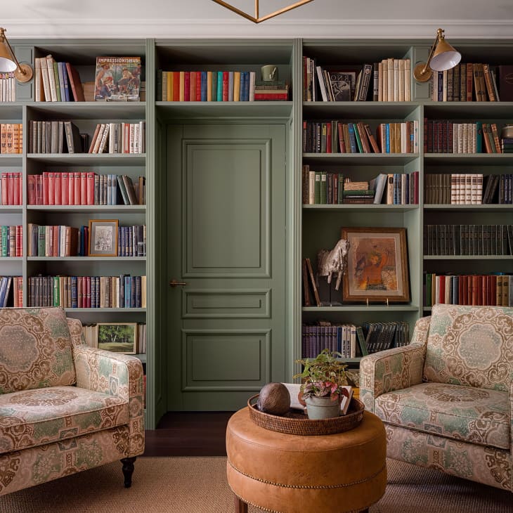 Sitting area with green wall-to-wall bookshelf, floral chairs, and round leather ottoman