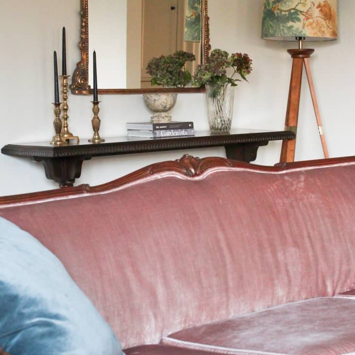 Vintage pink velvet sofa with blue cushions, ornate mirror, candlesticks, and floral decor on a wooden shelf.
