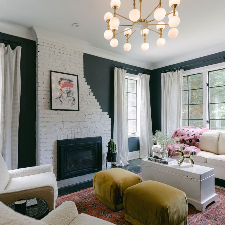 Living room with white brick fireplace, modern chandelier, cream sofa, mustard ottomans, and abstract artwork on dark walls.
