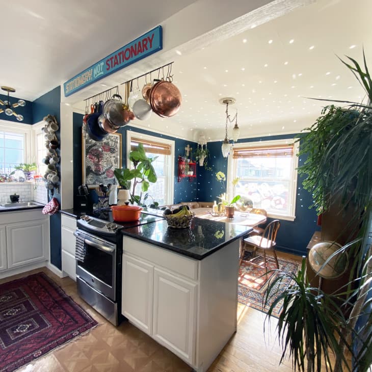 Kitchen with dark countertops, hanging pots, plants, and a dining nook with large windows and a patterned rug.