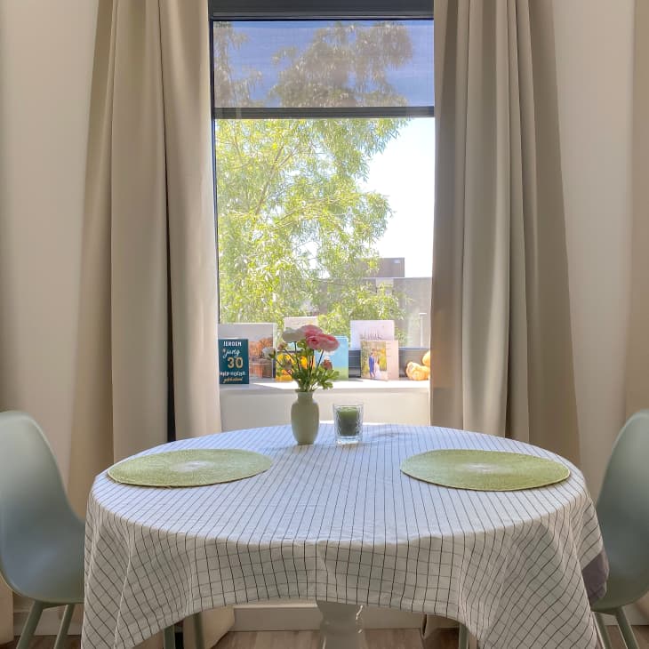 Round dining table with white cloth, two green placemats, and a vase of flowers by a window with beige curtains.
