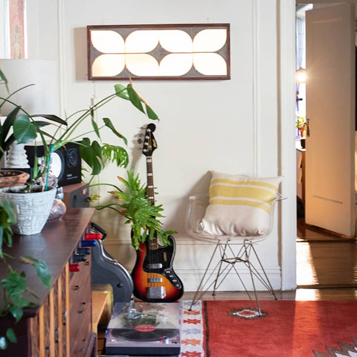Living room with plants, a guitar, record player, and a chair with a yellow-striped pillow, leading to an open door.
