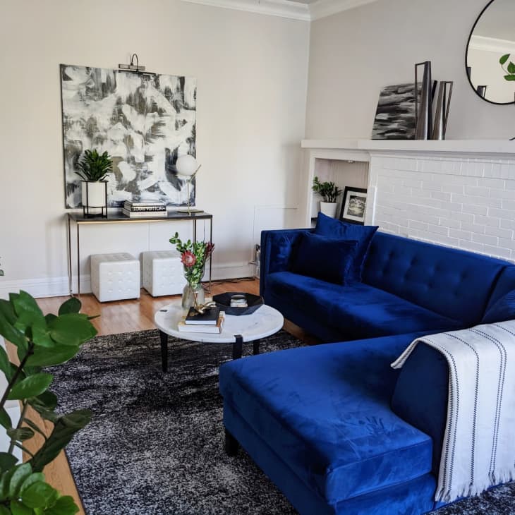 Living room with a blue velvet sectional sofa, white brick fireplace, abstract art, and plants on a console table.