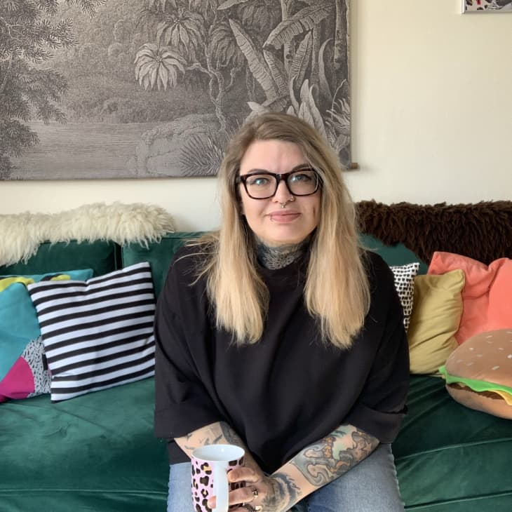 Woman with glasses and tattoos sitting on a green sofa with colorful pillows and a burger-shaped cushion.