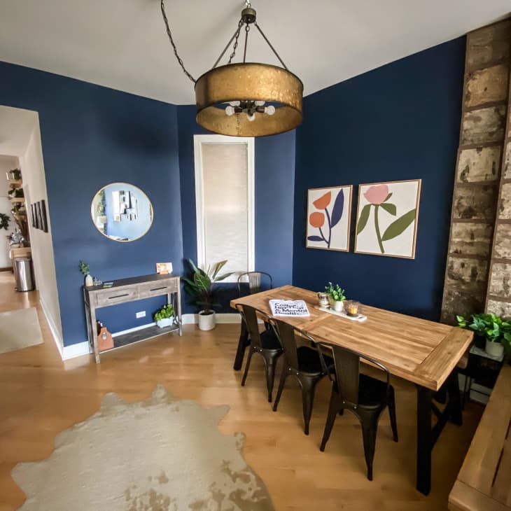 Dining room with blue walls, wooden table, black chairs, floral art, and a large pendant light.