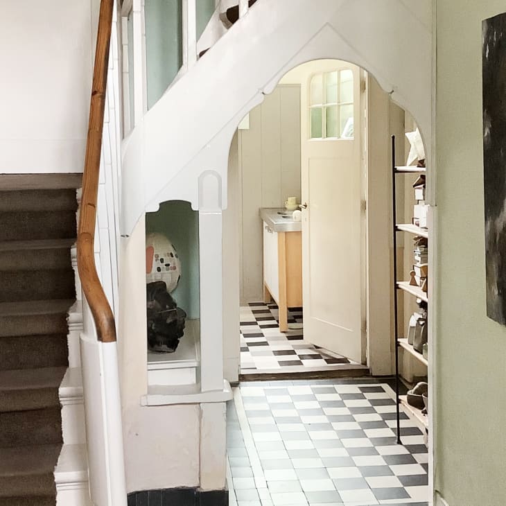 Staircase with brown carpet leads to an archway, revealing a kitchen with checkered floor and a painting on the wall.