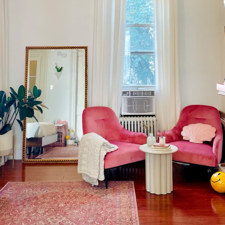 Two pink chairs with shell pillows, a small round table, and a large mirror in a cozy room with a plant and books on a mantel.
