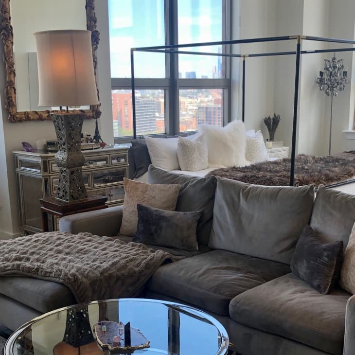 Living room with gray sectional sofa, mirrored dresser, canopy bed, and large windows overlooking cityscape.
