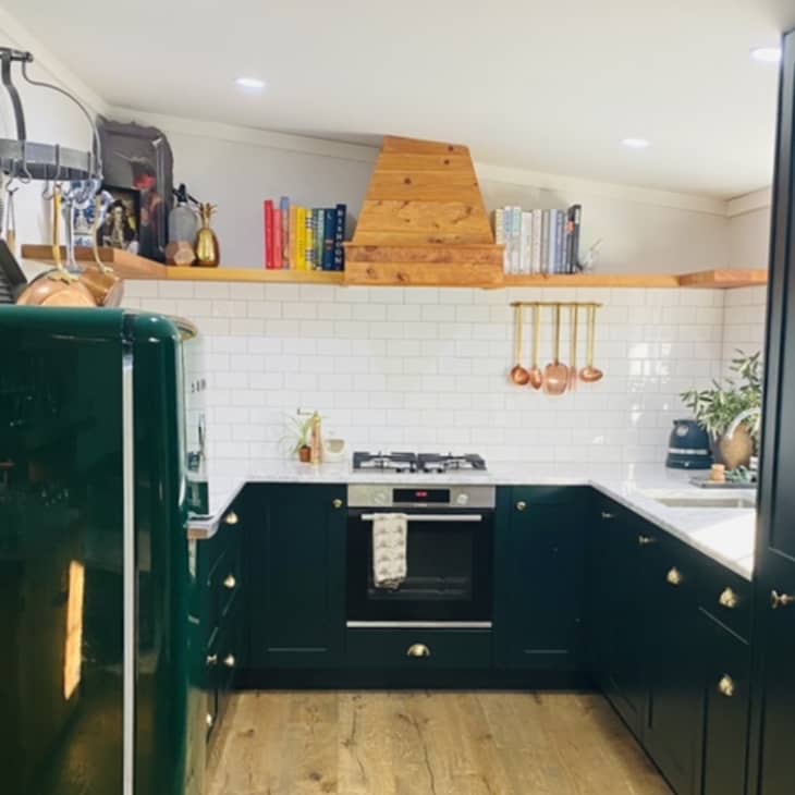 Dark green kitchen with wooden shelves, copper utensils, white subway tiles, and a vintage-style fridge.