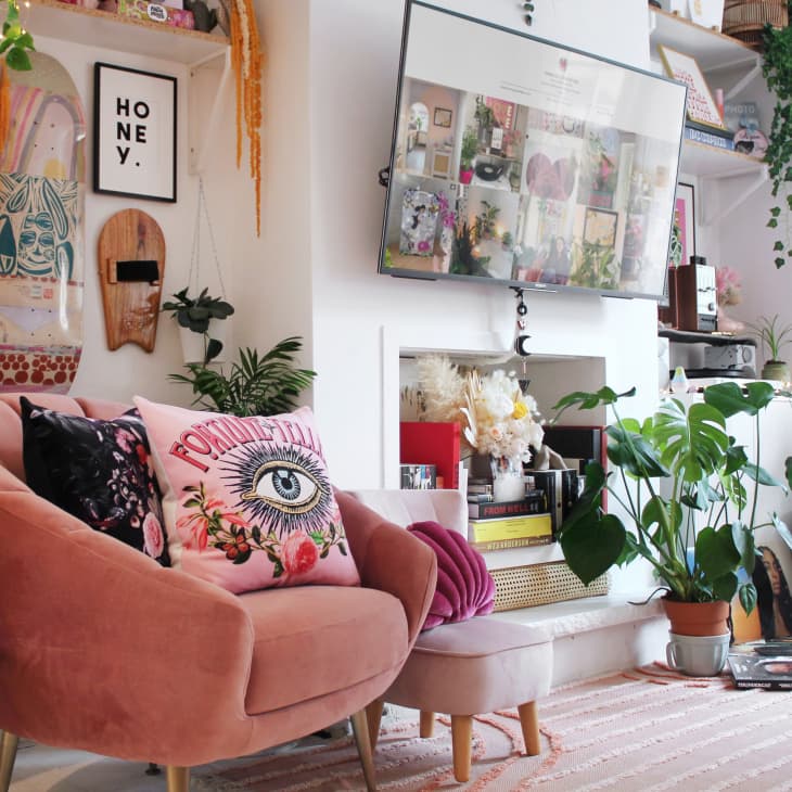 Cozy living room with pink armchair, eye-themed pillow, skateboard wall art, plants, and a wall-mounted TV.