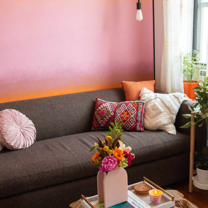 Cozy living room with a gray sofa, colorful pillows, pink wall, potted plants, and a vase of flowers on a coffee table.