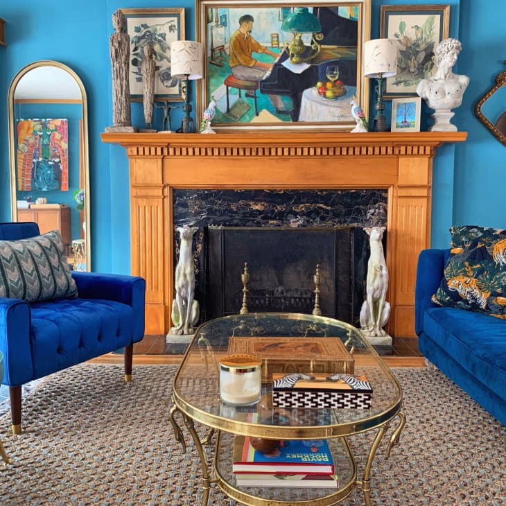 Blue living room with velvet chairs, patterned cushions, wooden fireplace, eclectic art, and a glass coffee table.
