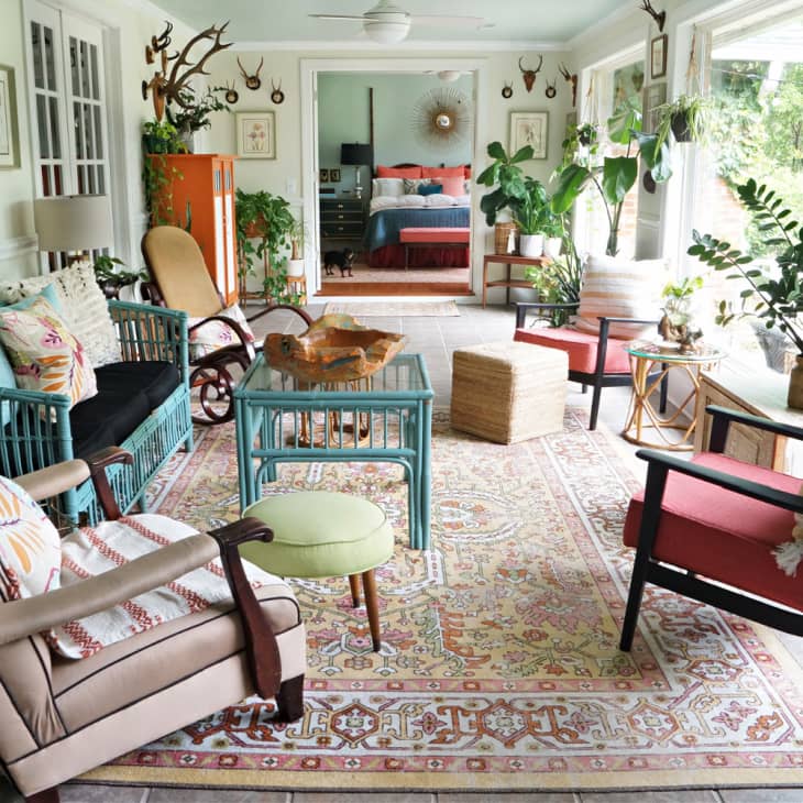 Eclectic sunroom with colorful furniture, patterned rug, plants, and antler wall decor leading to a bedroom.