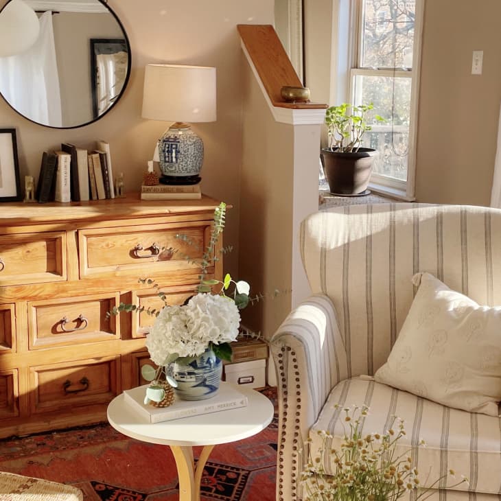 Cozy living room with striped armchair, wooden dresser, round mirror, and potted plants by a sunlit window.